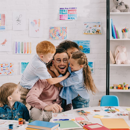 Image showing a nursery teacher sat at a desk being hugged by some of his class